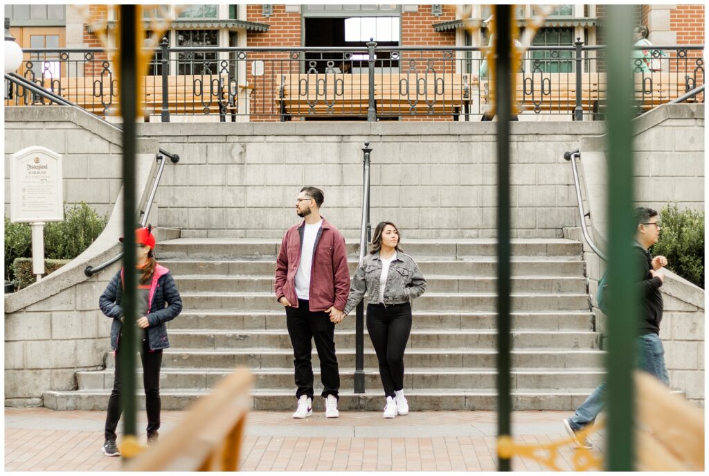 A couple stands hand in hand on a set of steps, with an urban backdrop of a brick building. They both look off into the distance, giving the image a candid, thoughtful feel.