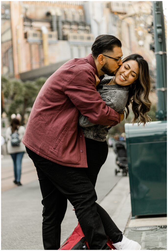 A couple shares a playful moment on a city street, with the man lifting and kissing the woman. They both smile, enjoying the lively urban surroundings.