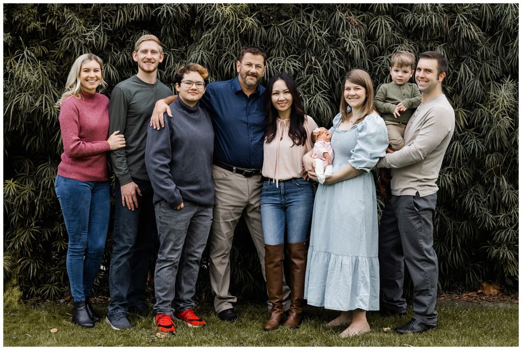 A family stands together, posing in front of a backdrop of dense, leafy plants. The group includes parents, young children, and other family members, all smiling at the camera.