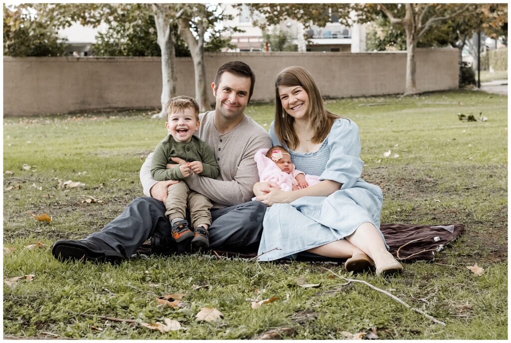 A young family sits on the grass, smiling at the camera. The parents hold their toddler son and newborn baby, creating a warm, familial scene in a park setting.