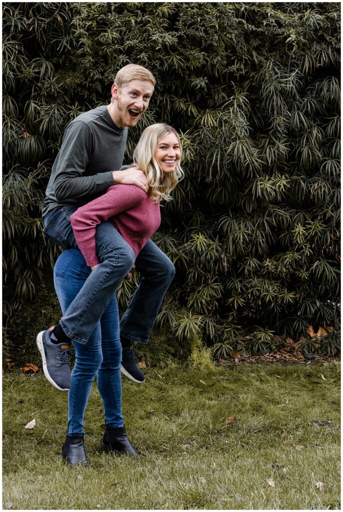 A man and woman share a playful moment as the woman gives the man a piggyback ride. They are both smiling brightly, standing on a grassy area with lush greenery in the background.