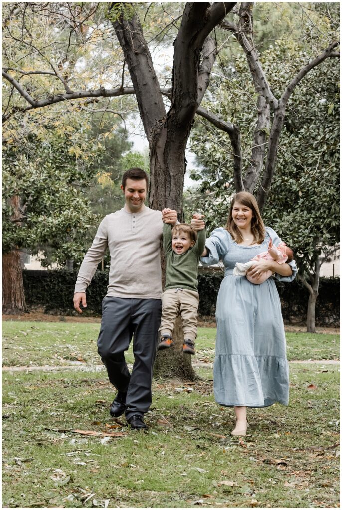 A family enjoys a walk in the park. The father and mother hold hands with their toddler son, lifting him playfully off the ground, while the mother also cradles a newborn baby. They are surrounded by trees and greenery.