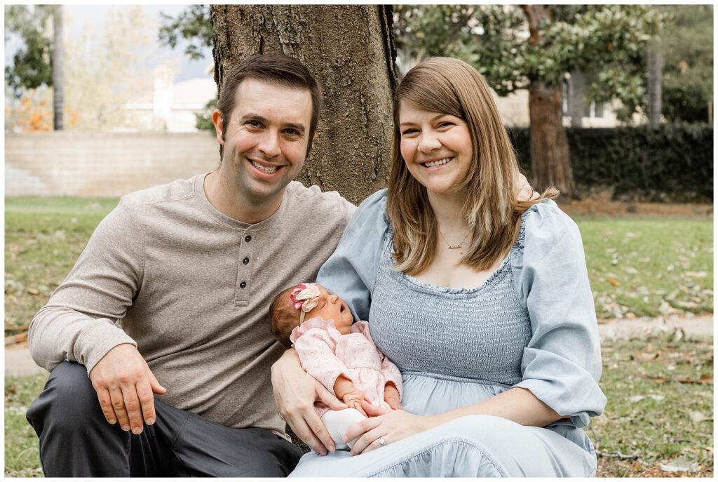 A couple sits by a tree in a park, holding their newborn baby. They are both smiling warmly at the camera, with the baby wrapped in a soft blanket.