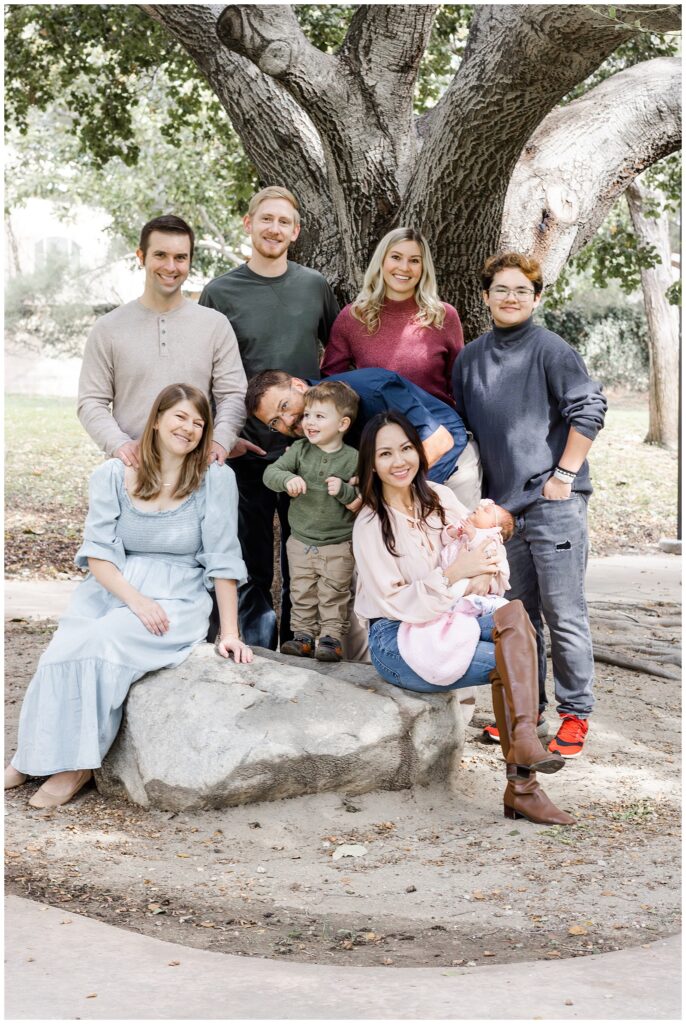 A large family group poses together in a park. They stand and sit around a large tree, smiling warmly at the camera. The group includes adults, a toddler, and a newborn baby.