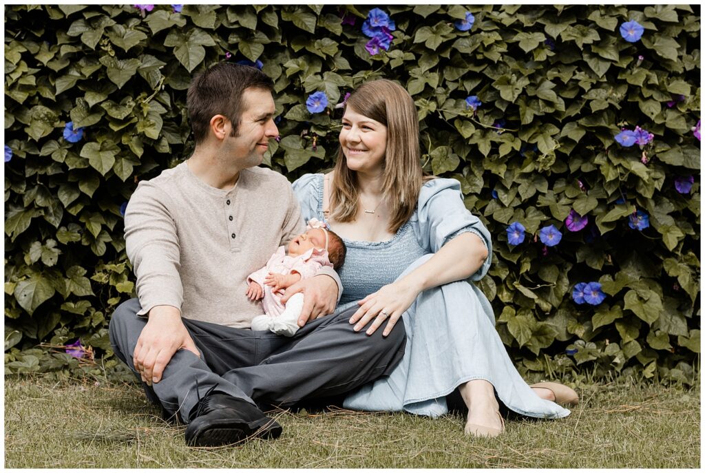 A couple sits on the grass in front of a flowering vine. They are smiling at each other, holding their newborn baby, creating a serene and loving family portrait.