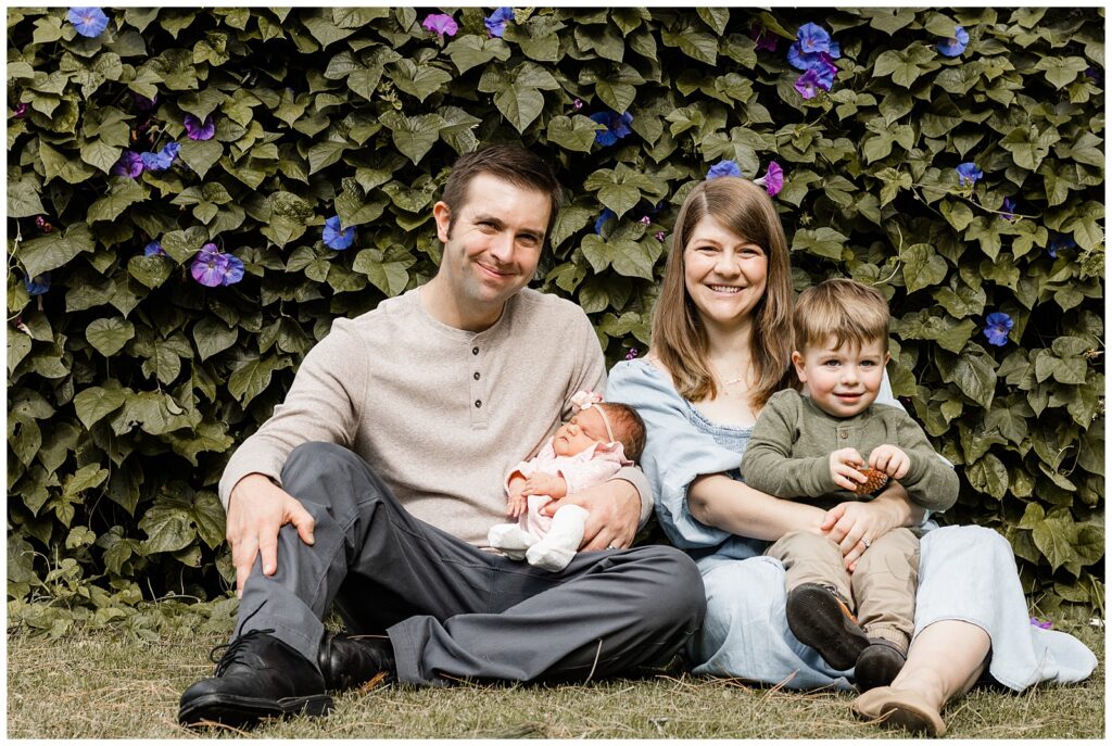 A family of four sits on the grass in front of a vibrant wall of flowers. The parents hold their newborn baby and toddler son, all smiling and enjoying the outdoor setting.