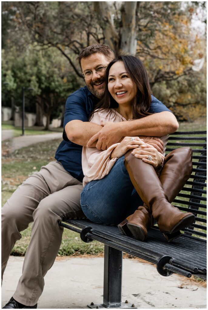 A couple sits closely together on a park bench. The man has his arms around the woman, and both are smiling contentedly. The park's natural scenery provides a serene backdrop.