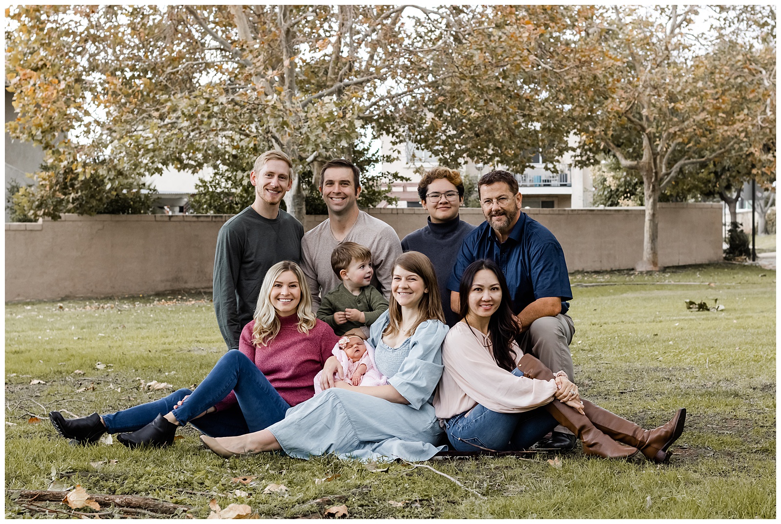 A large family group poses together on the grass in a park. They are seated and standing in a relaxed formation, smiling at the camera with trees and autumn foliage in the background.