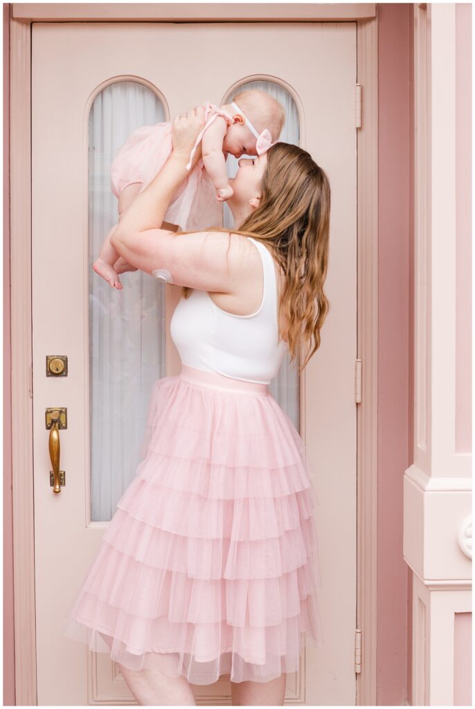 A woman in a white sleeveless top and a pink tulle skirt lifts a baby girl wearing a matching pink dress and bow headband. They are standing in front of a light pink door with oval windows. The woman is smiling at the baby, and they appear happy and joyful.