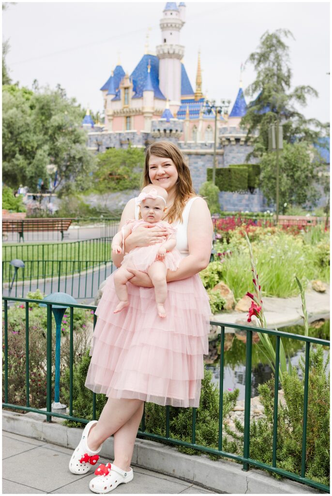 A woman in a white sleeveless top and a pink tulle skirt holds a baby girl wearing a matching pink dress and bow headband. They are standing outside with a view of a castle with blue rooftops in the background. The woman is smiling at the camera, and the baby looks curiously at the surroundings. The scene appears to be at a theme park, likely Disneyland.