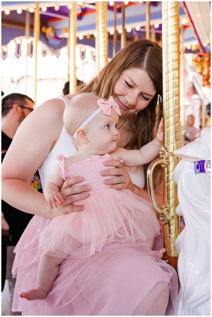 A woman in a white sleeveless top and a pink tulle skirt sits on a carousel, holding a baby girl wearing a matching pink dress and bow headband. The woman is looking affectionately at the baby, who is touching the gold-painted carousel pole. The background shows carousel lights and other riders, creating a festive and joyful atmosphere.