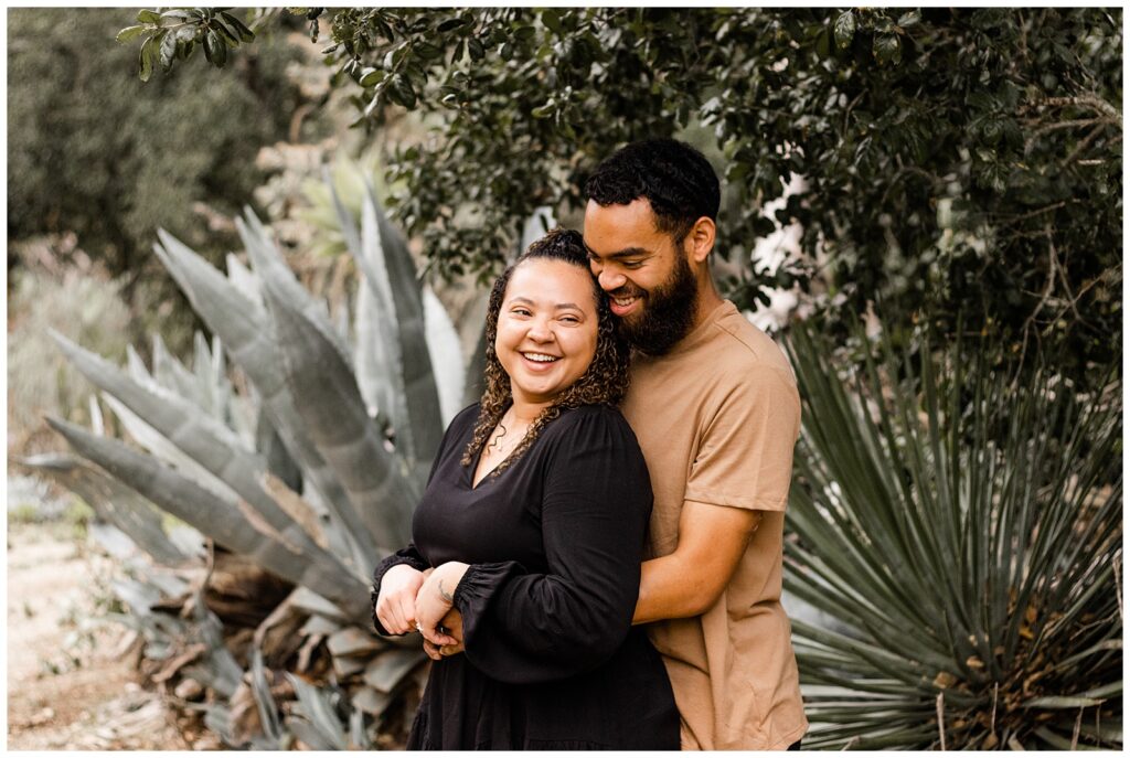 A couple standing close together, smiling and laughing, with the man standing behind the woman, holding her hands. They are outdoors with large agave plants and greenery in the background.