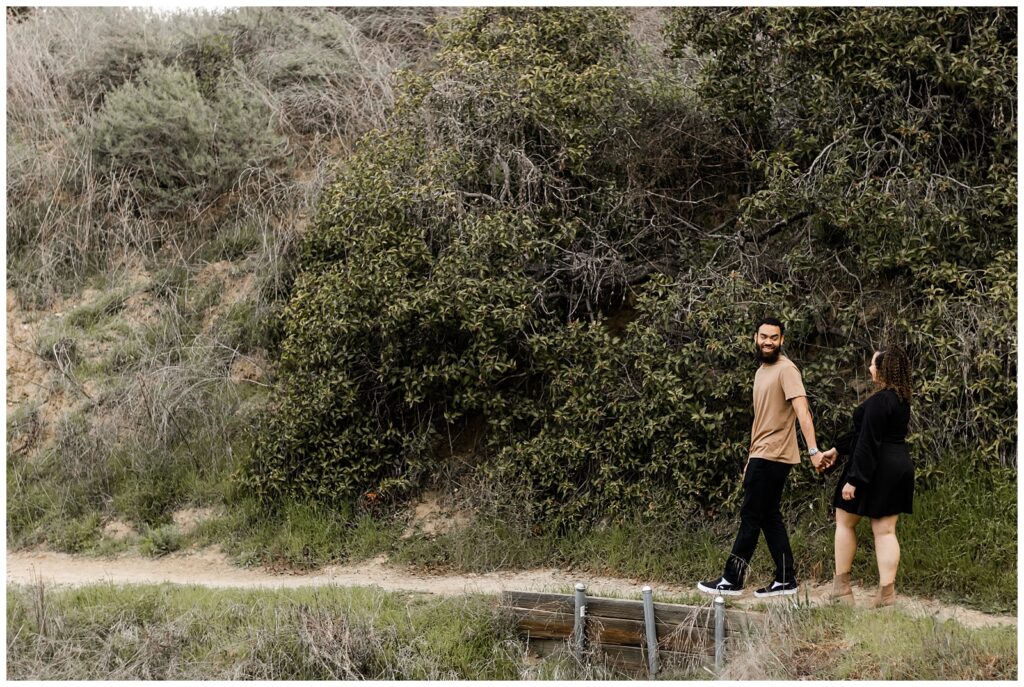 A couple holding hands and walking along a dirt path, surrounded by lush greenery and bushes. The man is looking back at the woman with a smile.