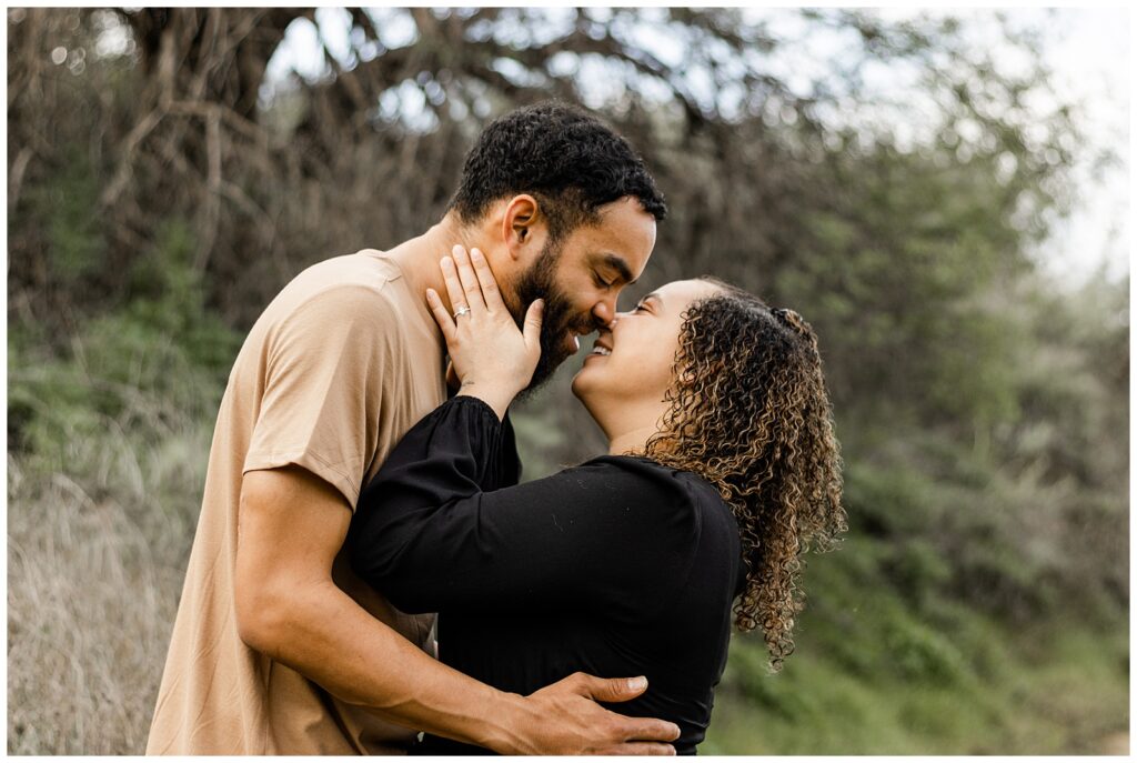 A couple standing face to face, about to kiss. The woman is gently holding the man's face, and they both have joyful expressions. The background is a natural, outdoor setting with trees and shrubs.