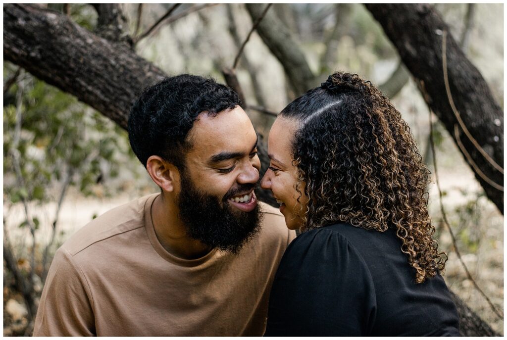 A couple sitting close together, leaning in with their noses touching, both smiling and sharing a tender moment. They are surrounded by tree branches and foliage.