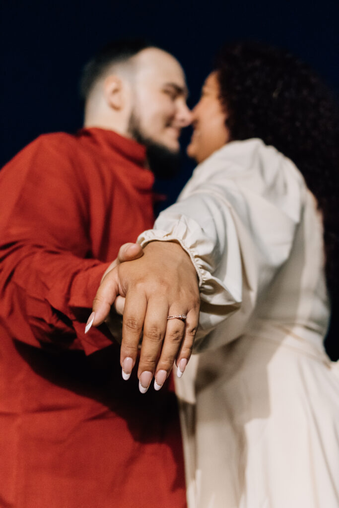 A couple is shown in an artistic shot, with the focus on the woman's engagement ring as she holds her partner's hand. Both are slightly out of focus as they lean in for a kiss in the background, creating a dreamy and romantic image.