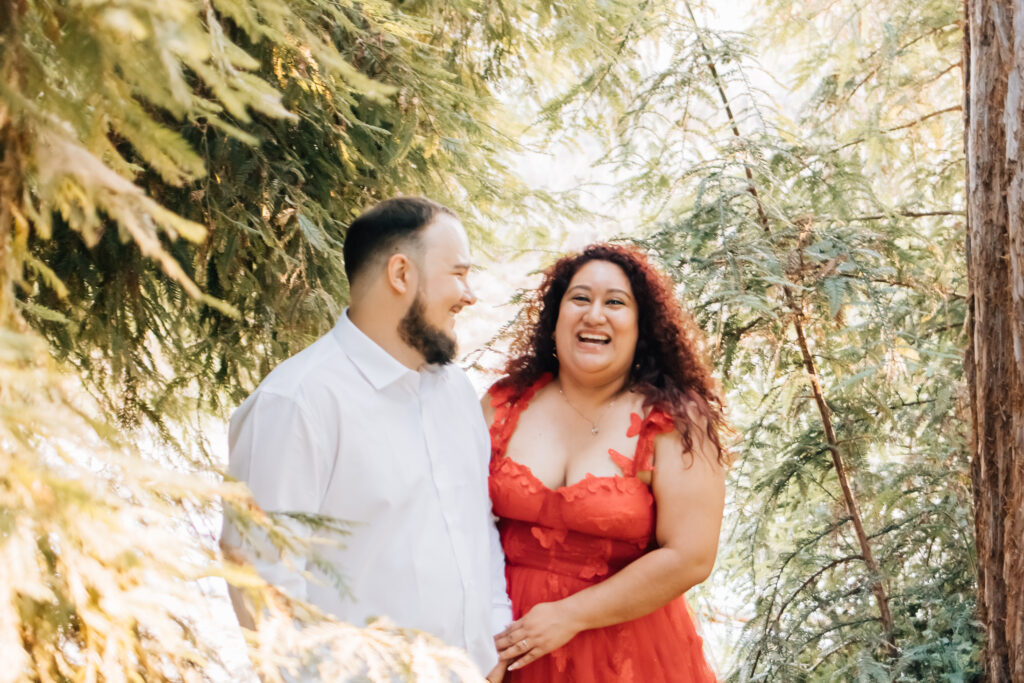 A joyful couple stands together in a forest, bathed in warm sunlight filtering through the trees. The woman, wearing a vibrant red dress, laughs openly while looking at the man, who is smiling at her. The background features lush greenery, creating a serene and romantic atmosphere.