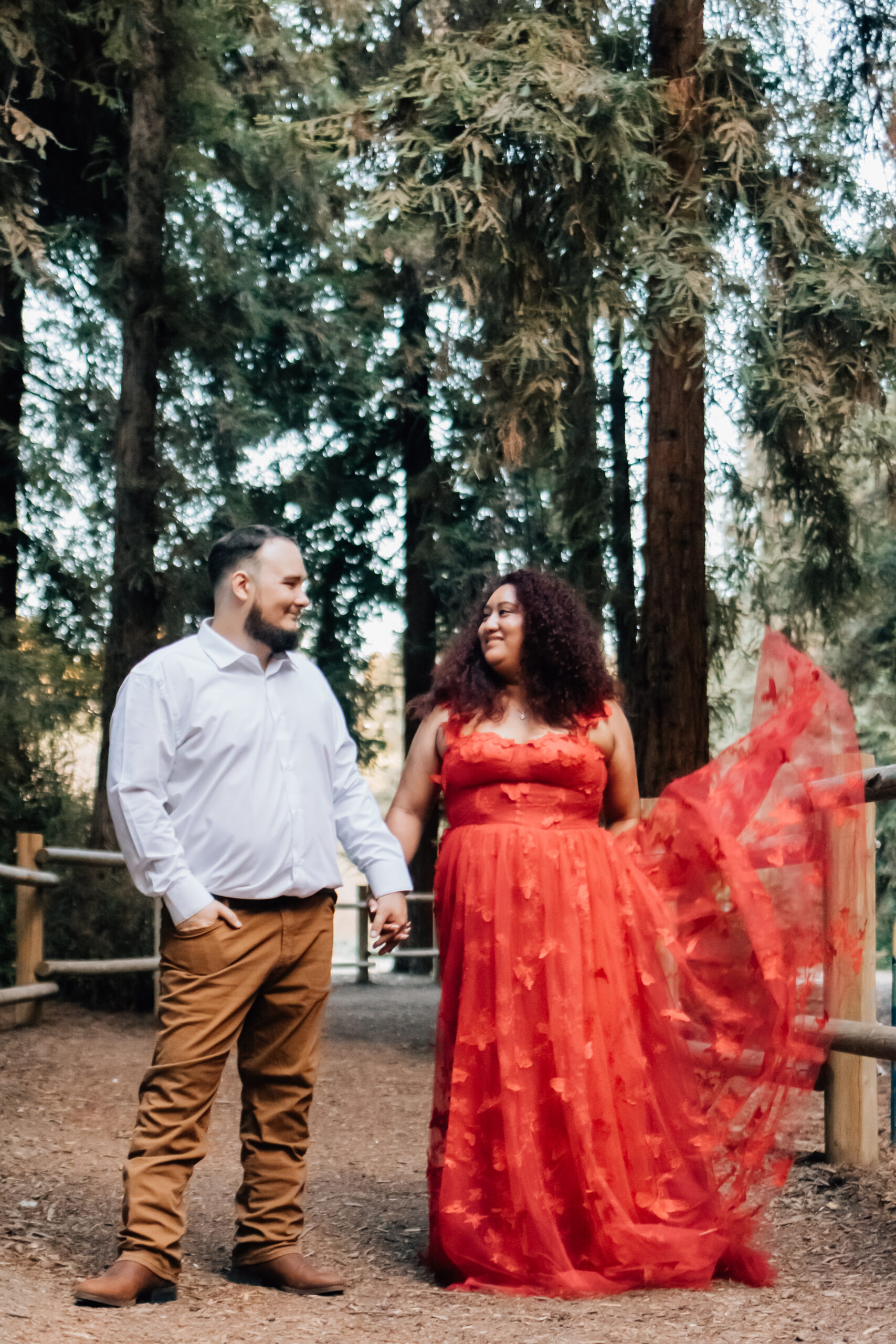 A couple walks hand in hand through a wooded area, with the woman's red dress flowing behind her. They look at each other warmly, sharing a quiet moment in the serene forest surroundings.