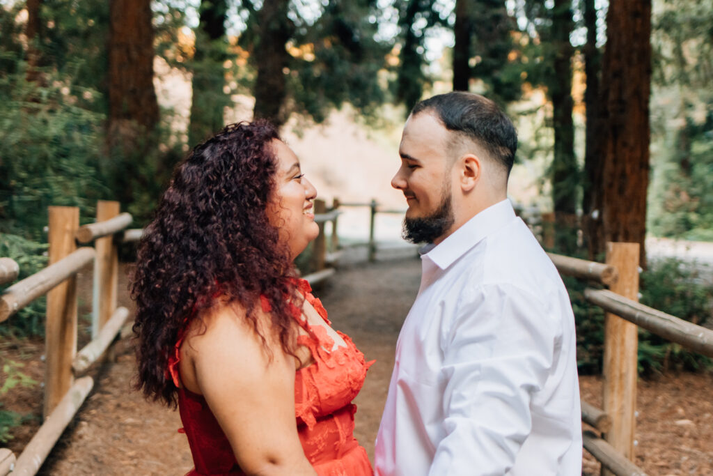 A couple stands facing each other on a path lined with wooden railings, surrounded by tall trees. They are smiling at each other, creating an intimate and loving moment captured in nature.