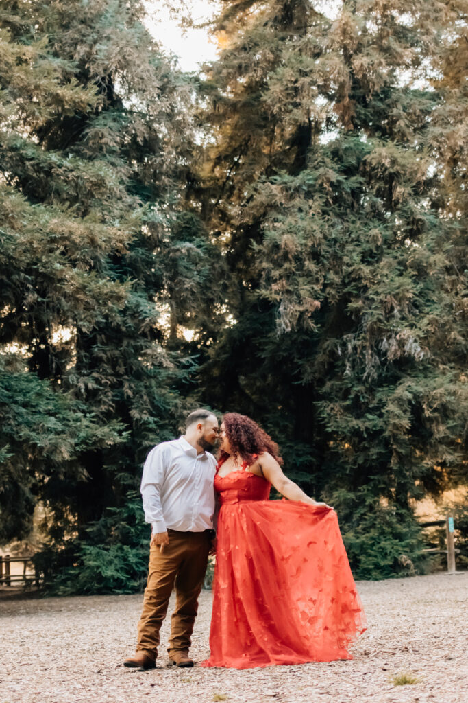 A couple stands close together in front of a large group of tall trees. The woman is dressed in a red dress, and the man in a white shirt and brown pants. The woman holds the hem of her dress as the man leans in to kiss her forehead, creating a romantic and serene scene.