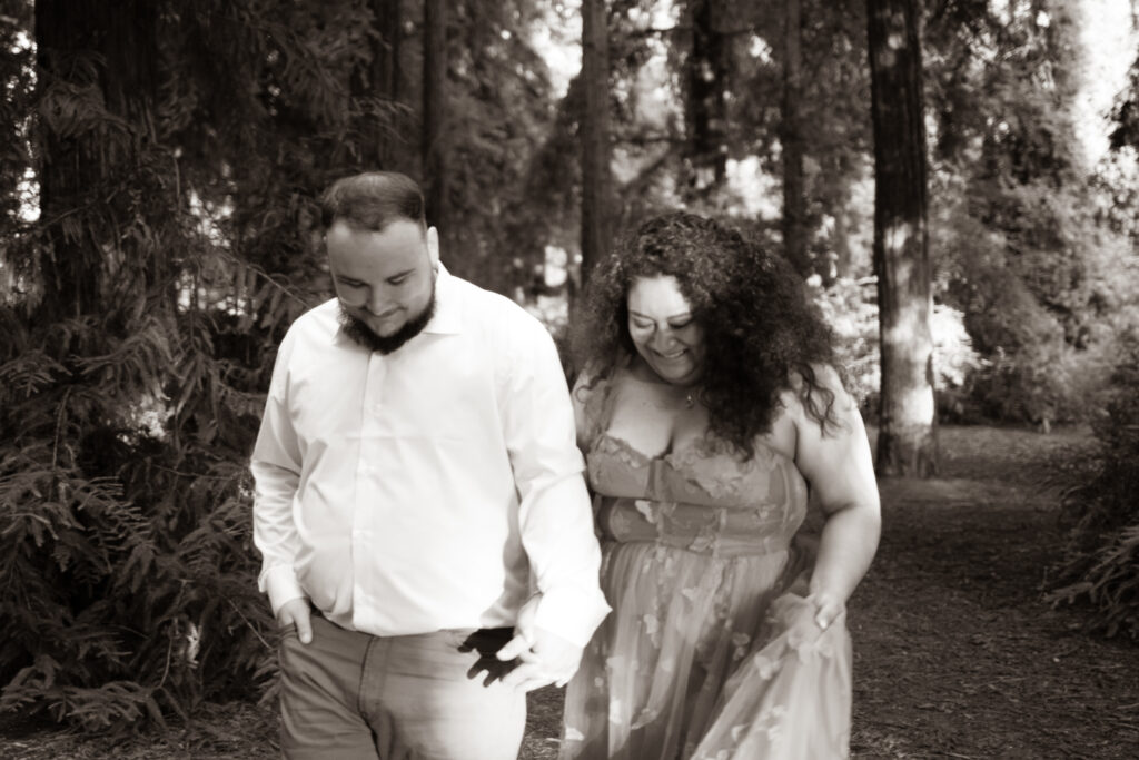 A black-and-white photo of a couple walking together through a forest. They are both looking down and smiling, capturing a candid moment of joy as they hold hands. The trees in the background provide a soft, textured backdrop.