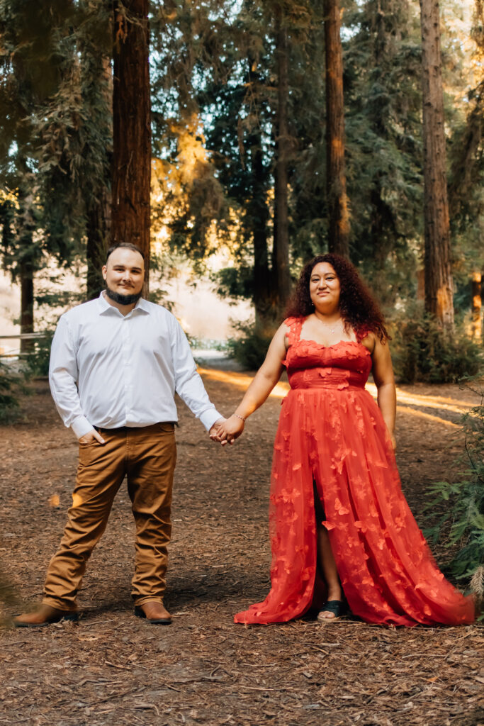 A couple stands hand in hand on a forest path, surrounded by tall trees. The woman is wearing a striking red dress, and the man is dressed in a white shirt and brown pants. They both look into the camera with calm expressions, framed by the natural beauty of the forest.