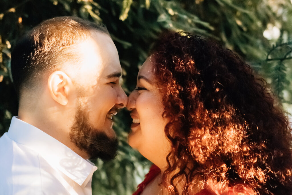 A close-up image of a couple smiling as they stand nose-to-nose in a forest. The sunlight gently highlights the woman's curly hair and the man's face, capturing a tender and happy moment between them.