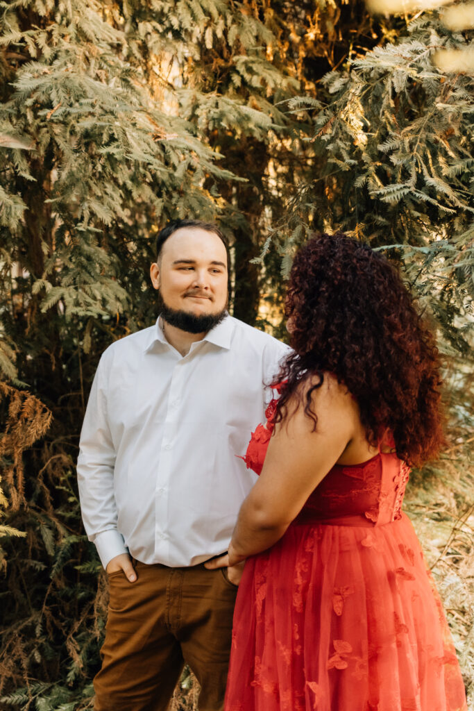 A man and a woman stand closely together in a forest setting. The man, dressed in a white shirt, looks thoughtfully at the woman, who is wearing a flowing red dress. The natural green backdrop provides a calm and intimate environment for the couple's portrait.