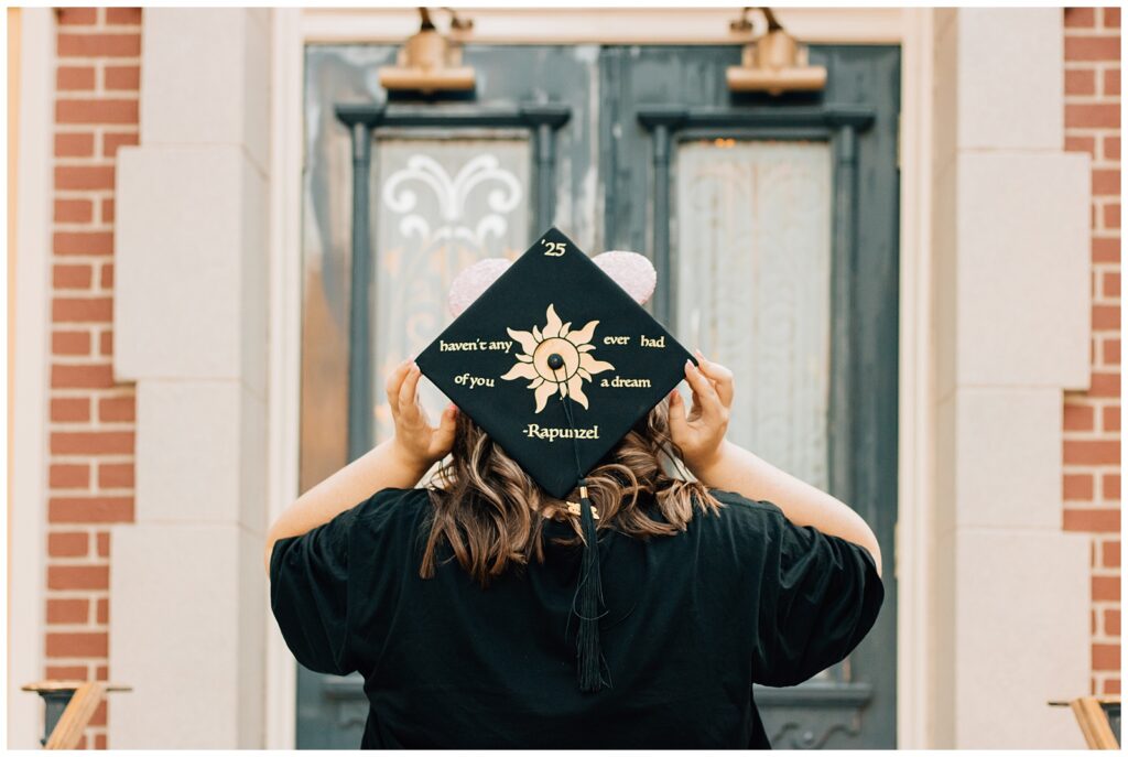 A young woman holds her graduation cap behind her head, displaying the Tangled-inspired design that reads, "Haven’t any of you ever had a dream?" with the Rapunzel sun symbol. She stands in front of a detailed doorway in the park.