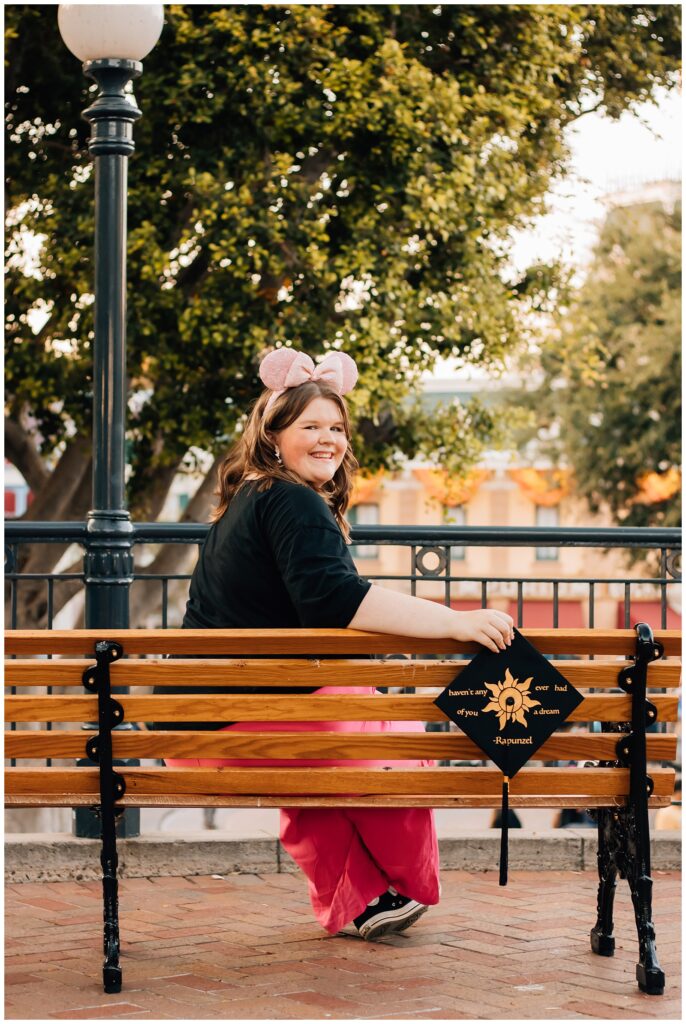 A young woman, wearing pink mouse ears and holding her graduation cap, sits on a bench in a Disney park, smiling at the camera, with trees and park scenery in the background.