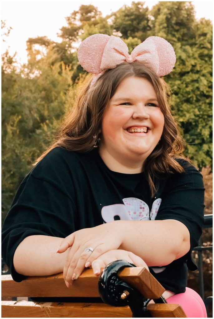 Close-up of a young woman sitting on a bench at a Disney park, smiling warmly with sparkly pink mouse ears on her head, enjoying the ambiance of the park.