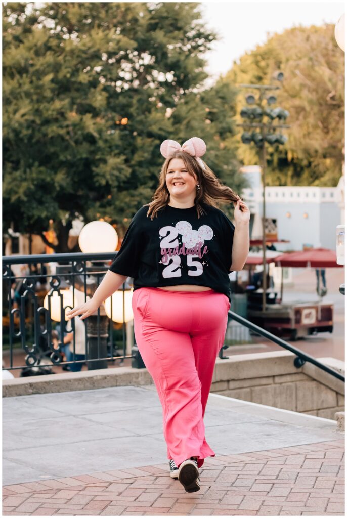 A joyful young woman wearing bright pink pants, a 2025 graduation t-shirt, and pink mouse ears walks confidently at a Disney park, her hair flowing in the breeze.