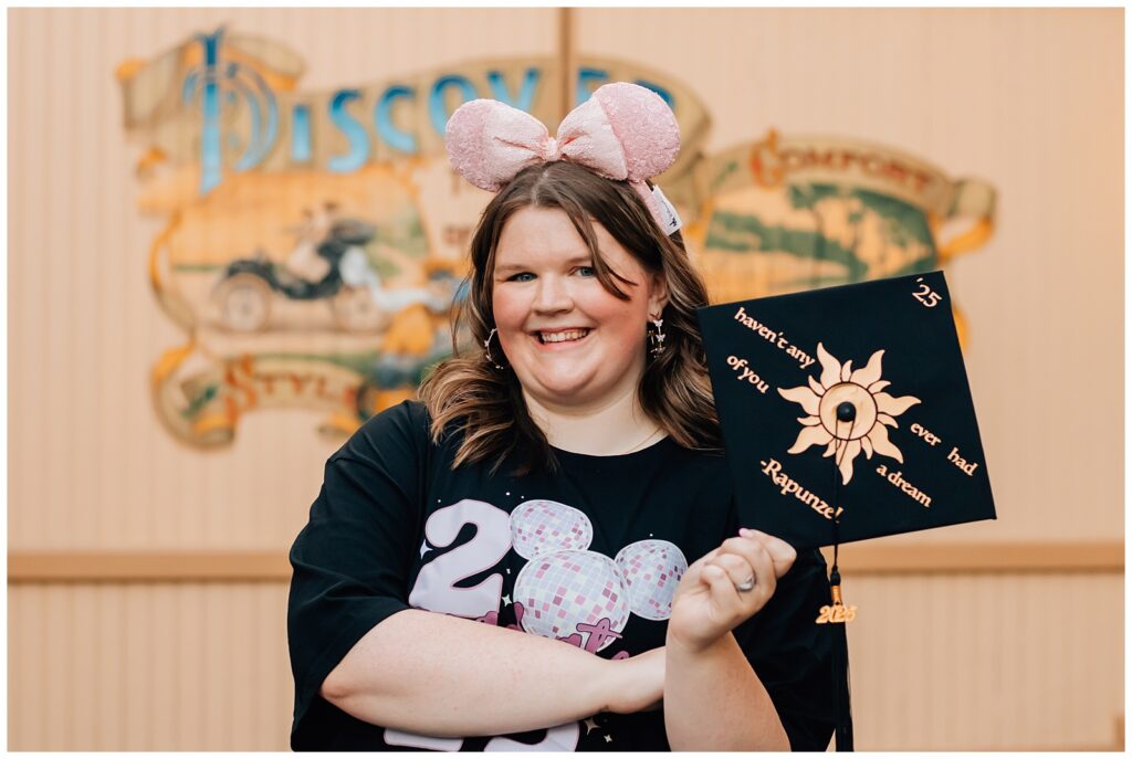 A young woman stands proudly at a Disney park, holding a graduation cap decorated with the sun symbol from Tangled and a quote that reads, "Haven’t any of you ever had a dream? –Rapunzel."