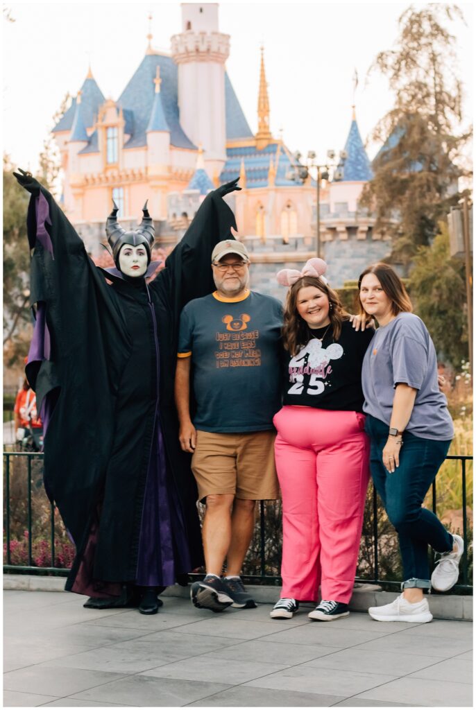 A group photo at a Disney park, featuring a young woman in pink pants and pink mouse ears, standing with her family and a costumed Maleficent character, with the iconic Disney castle in the background.