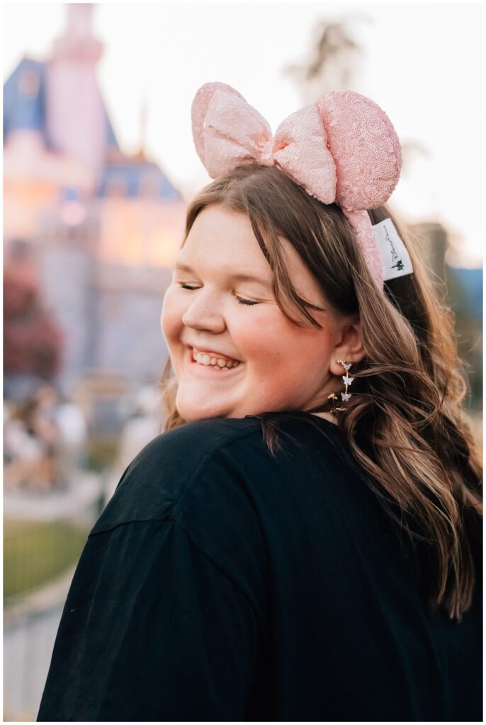 A smiling young woman wears sparkly pink mouse ears while standing in front of a castle at a Disney park. Her eyes are closed, and she looks over her shoulder, exuding happiness.