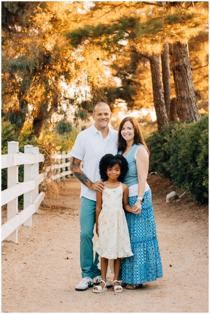 A family of three posing together on a dirt pathway surrounded by greenery. The father, with tattoos and wearing a white shirt and teal pants, stands to the left. The mother, in a blue vest and patterned skirt, stands to the right, while their daughter, in a light floral dress, stands in front of them, smiling.