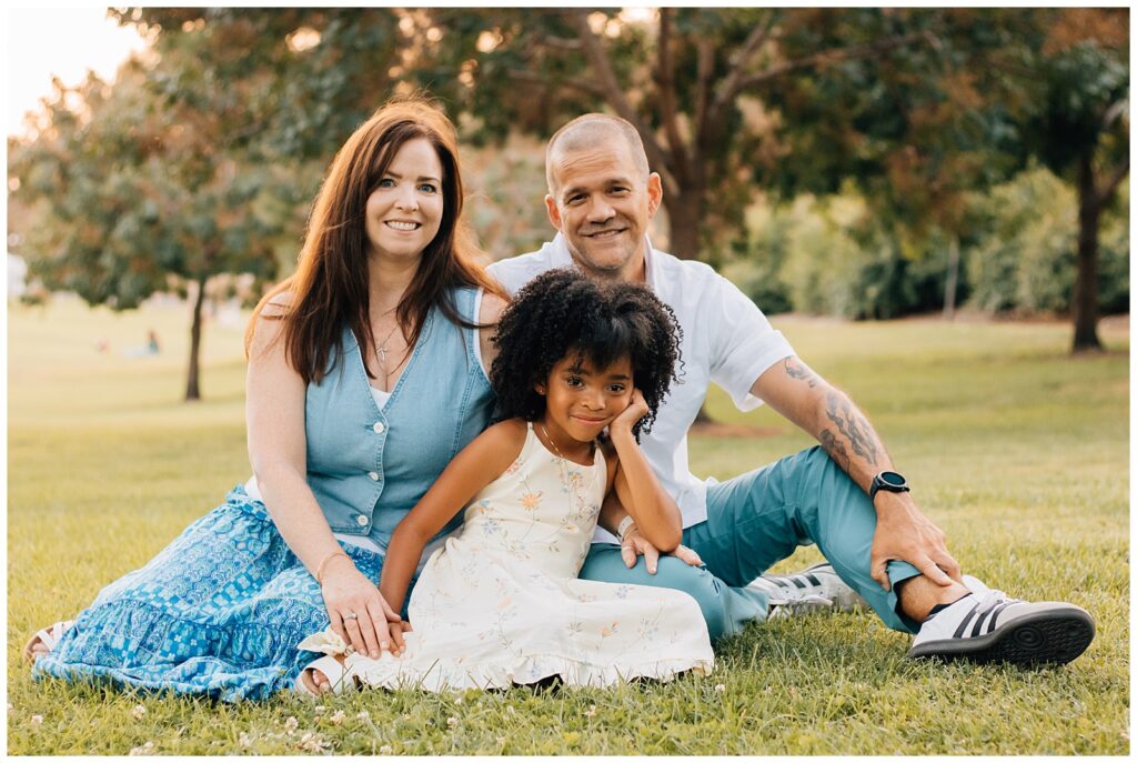 The family sits together on the grass in a park, smiling at the camera. The mother and father sit cross-legged, with their daughter sitting in front of them, resting her head on her hand. The family appears relaxed and happy, enjoying the outdoor setting during sunset.