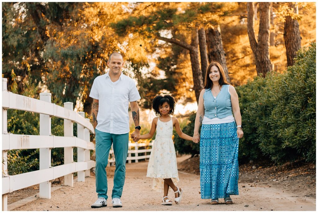 The family of three walking hand in hand along a rustic dirt path with trees in the background. The daughter, wearing a floral dress, is in the middle holding her parents' hands. The parents are smiling as they walk alongside a white fence.