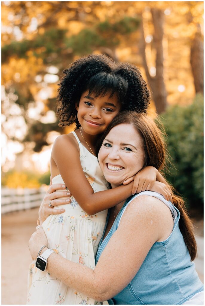 A close-up shot of the mother and daughter embracing. The daughter, in a floral dress, wraps her arms around her mother's neck, while the mother smiles warmly. They are outdoors with soft sunlight filtering through the trees behind them.