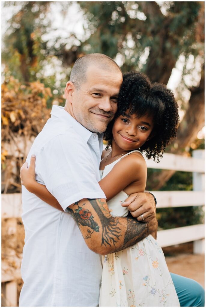 A close-up photo of the father and daughter hugging. The father, with visible tattoos, wears a white shirt, while the daughter, in a floral dress, smiles sweetly at the camera. The background features a blurred, natural outdoor setting.