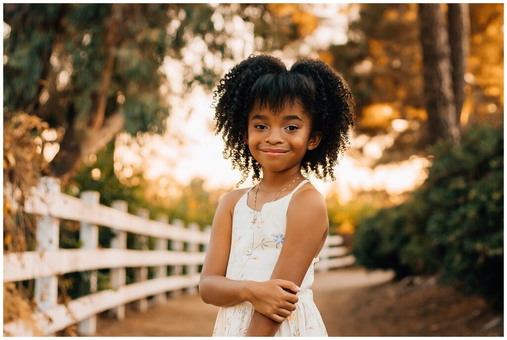 A solo portrait of the young girl standing with her arms crossed, wearing a floral dress. She smiles confidently as she stands on a pathway with trees and a white fence in the background, bathed in warm sunlight.