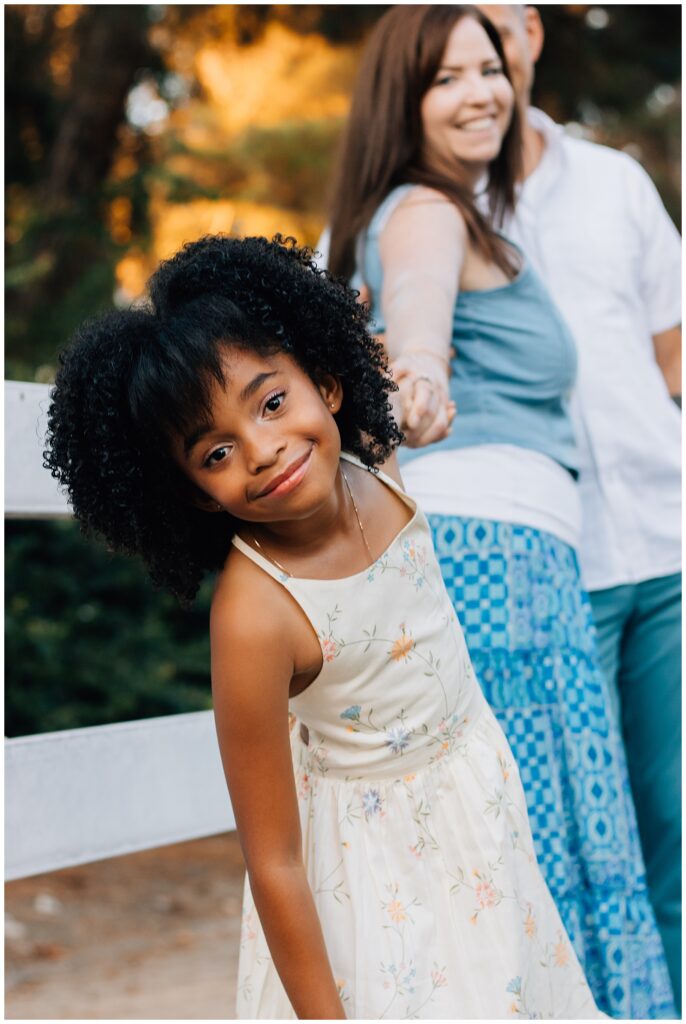 The young girl leans playfully toward the camera, smiling brightly as her parents stand behind her holding hands. The mother wears a long blue skirt, and the father sports teal pants. They are in a park-like setting with soft, golden light from the setting sun.