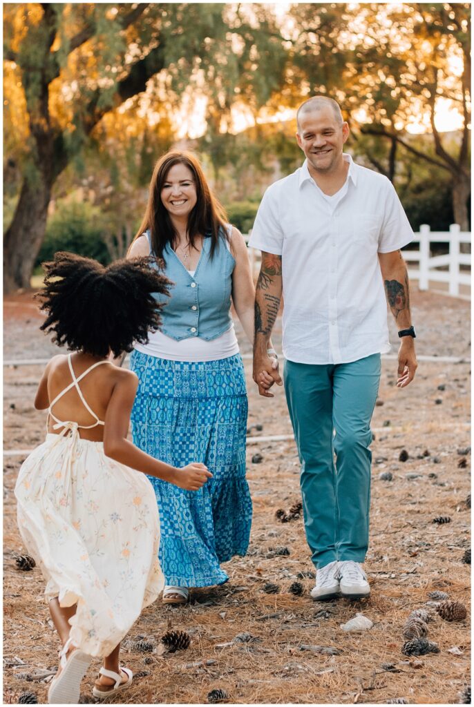 A candid shot of the parents walking hand in hand as their daughter runs toward them, her back to the camera. The family is walking through an open, wooded area, with pinecones scattered on the ground.