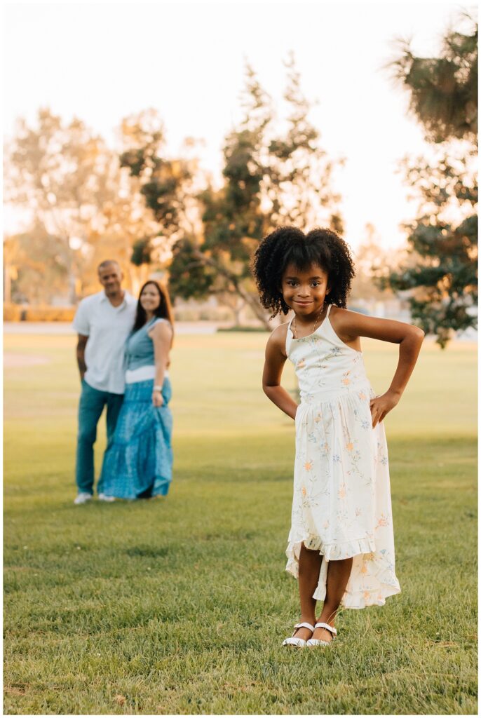 The young girl stands in the foreground with her hands on her hips, smiling playfully, while her parents stand in the background, slightly out of focus, in a grassy park setting during golden hour.