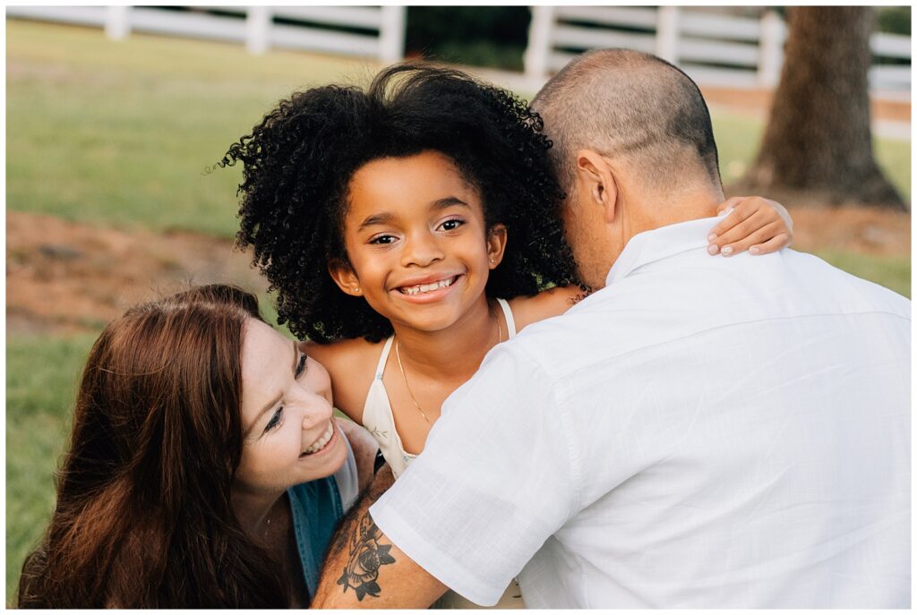 A close-up of the family gathered together in a group hug. The daughter smiles joyfully toward the camera, with her parents' faces partially visible as they embrace her lovingly. The image is filled with warmth and laughter.