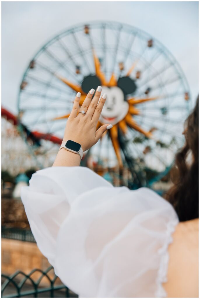 A hand with a silver engagement ring raised in front of the Mickey's Fun Wheel at Disneyland's California Adventure, symbolizing a special engagement moment. The wearer is dressed in a white, puffy-sleeved dress.