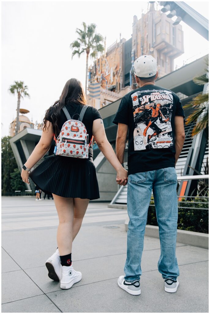 A couple holding hands, walking away from the camera, with the Avengers Campus backdrop at Disneyland’s California Adventure. The woman’s Marvel-themed backpack and skirt complement the man’s Spider-Man-themed shirt as they head toward the Guardians of the Galaxy ride.