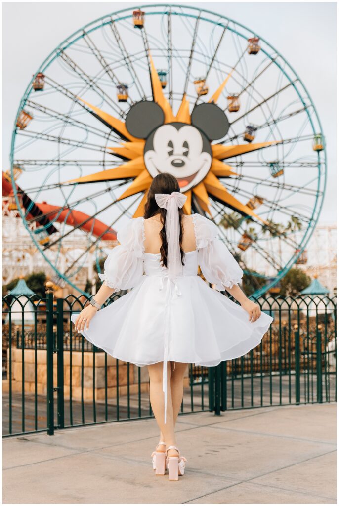 A woman wearing a white, puffy-sleeved dress with a large bow on the back stands in front of Mickey's Fun Wheel at Disneyland's California Adventure. Her back is facing the camera as she embraces the whimsical moment.