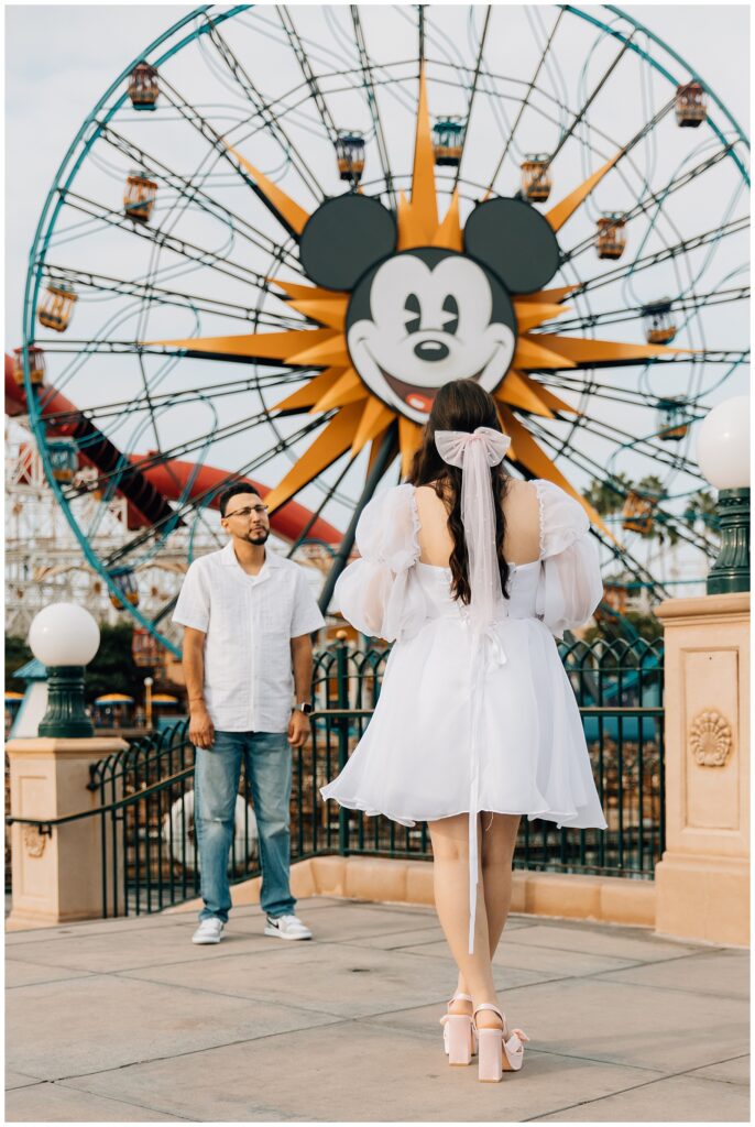 The woman in a white dress faces Mickey's Fun Wheel at Disneyland's California Adventure, while her partner stands in front of her, gazing at her with a loving expression.