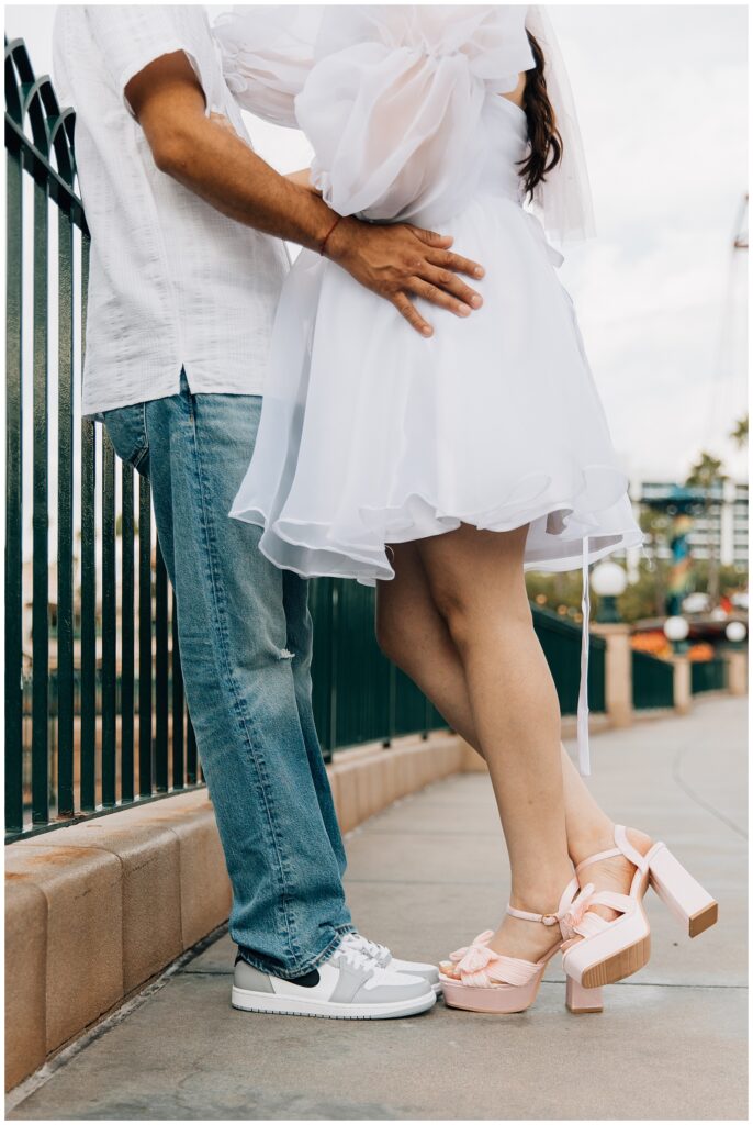 A close-up shot of a couple’s hands. The woman is wearing pastel pink platform heels, while the man is wearing casual white and gray sneakers. They are standing close, embracing in front of a Disneyland California Adventure fence.